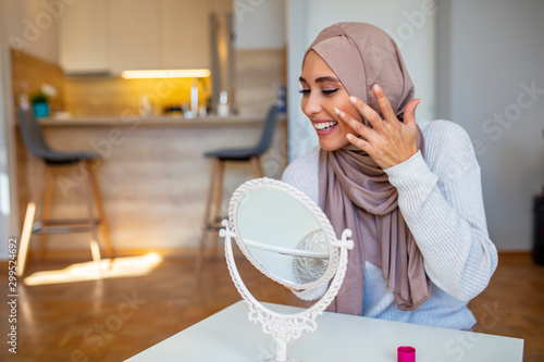 Smiling Muslim woman looking into hand mirror and applying face cream. Young Muslim woman looking at mirror at home and applying cream on her face. Let's put some cream on
