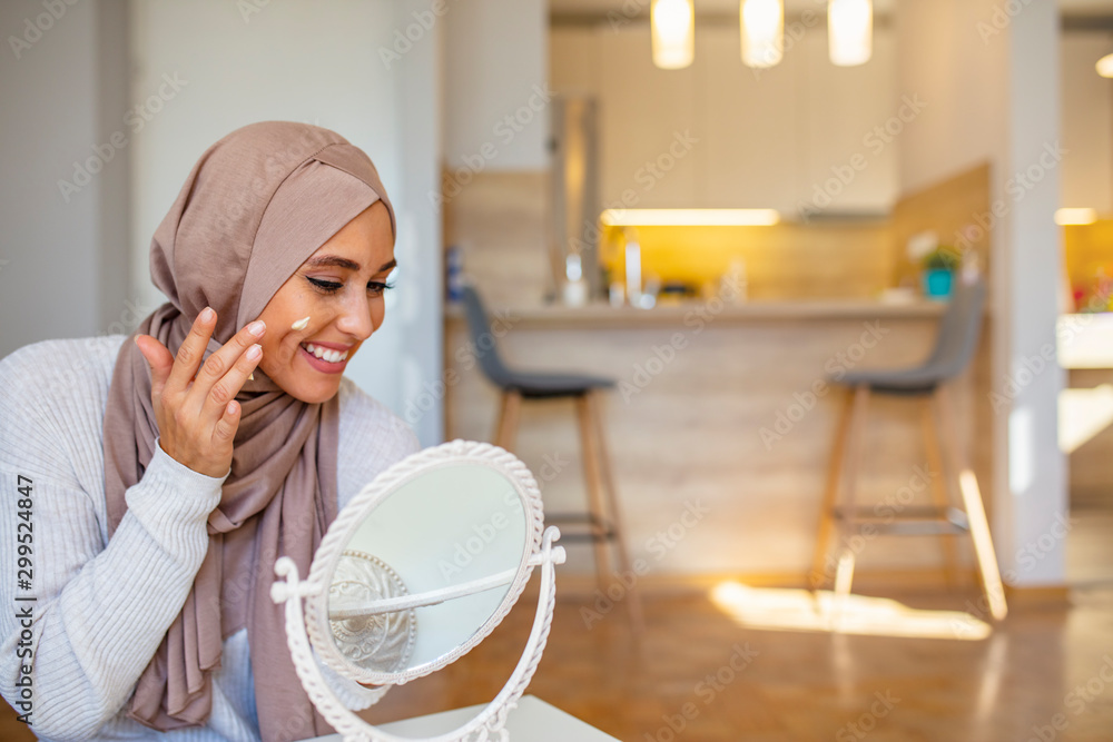 Muslim woman spreading cream over her face while looking in the mirror. Beauty treatment. Female putting on moisturizer on her facial. Smiling Muslim woman applying cream to face and looking to mirror Muslim woman spreading cream over her face while looking in the mirror. Beauty treatment. Female putting on moisturizer on her facial. Smiling Muslim woman applying cream to face and looking to mirror
