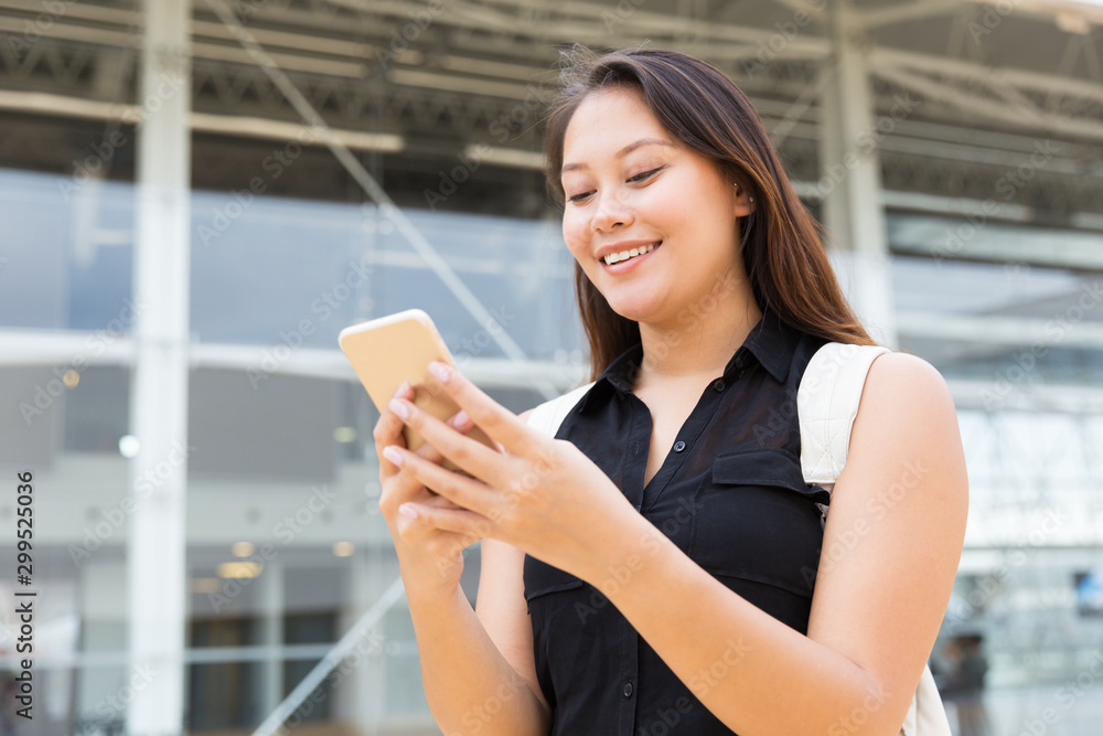 Positive female student texting message on cell. Young mix raced woman standing outside, using mobile phone, looking at screen, smiling. Digital technology concept Positive female student texting message on cell. Young mix raced woman standing outside, using mobile phone, looking at screen, smiling. Digital technology concept