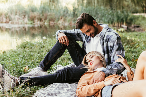 handsome man embracing girlfriend sleeping on blanket near lake in park