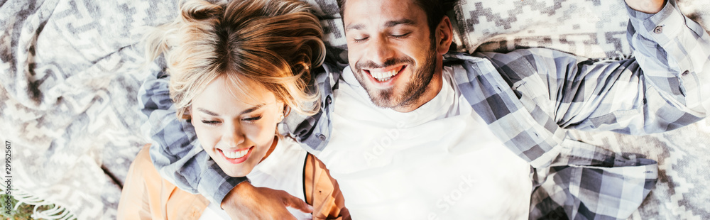 top view of cheerful man embracing happy girlfriend while lying on blanket in park, panoramic shot - obrazy, fototapety, plakaty 