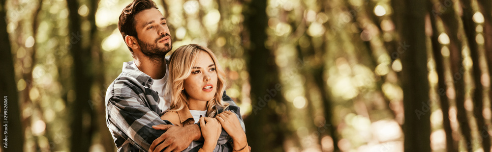 Fototapeta premium panoramic shot of happy man embracing attractive girlfriend while looking away in park