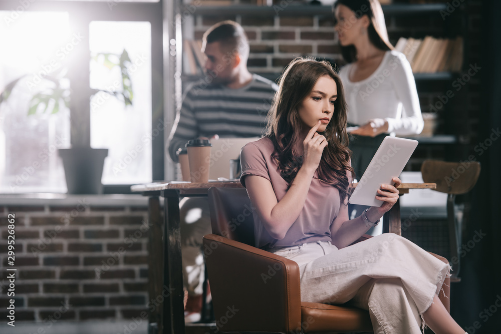 Fototapeta premium thoughtful businesswoman sitting in armchair and using digital tablet near young colleagues