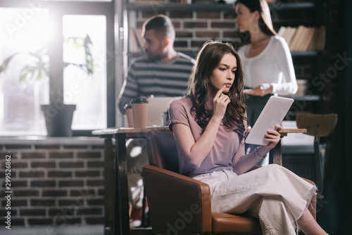 thoughtful businesswoman sitting in armchair and using digital tablet near young colleagues