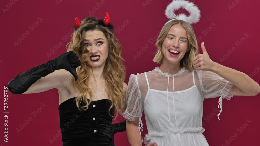 Sad demon girl shows dislike gestures while happy angel girl shows like gestures and they looking at the camera in carnival costumes isolated over red wall background Sad demon girl shows dislike gestures while happy angel girl shows like gestures and they looking at the camera in carnival costumes isolated over red wall background