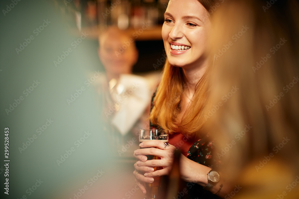 Portrait of cheerful caucasian blonde standing in pub, holding pint of beer and chatting with friends. Nightlife.