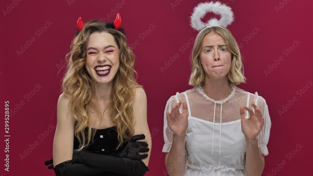 Happy demon girl incites to action and the sad angel girl dissuades them in carnival costumes isolated over red wall background Happy demon girl incites to action and the sad angel girl dissuades them in carnival costumes isolated over red wall background