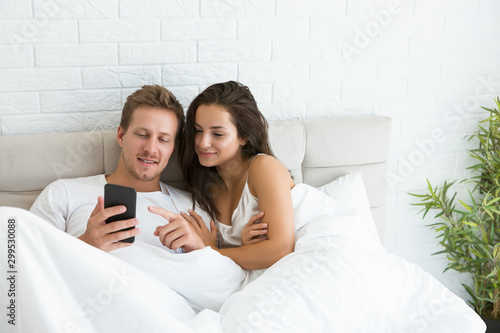 young couple handsome husband and his beautiful wife just woke up in bed watching photoes on smartphone early in the morning social media addiction