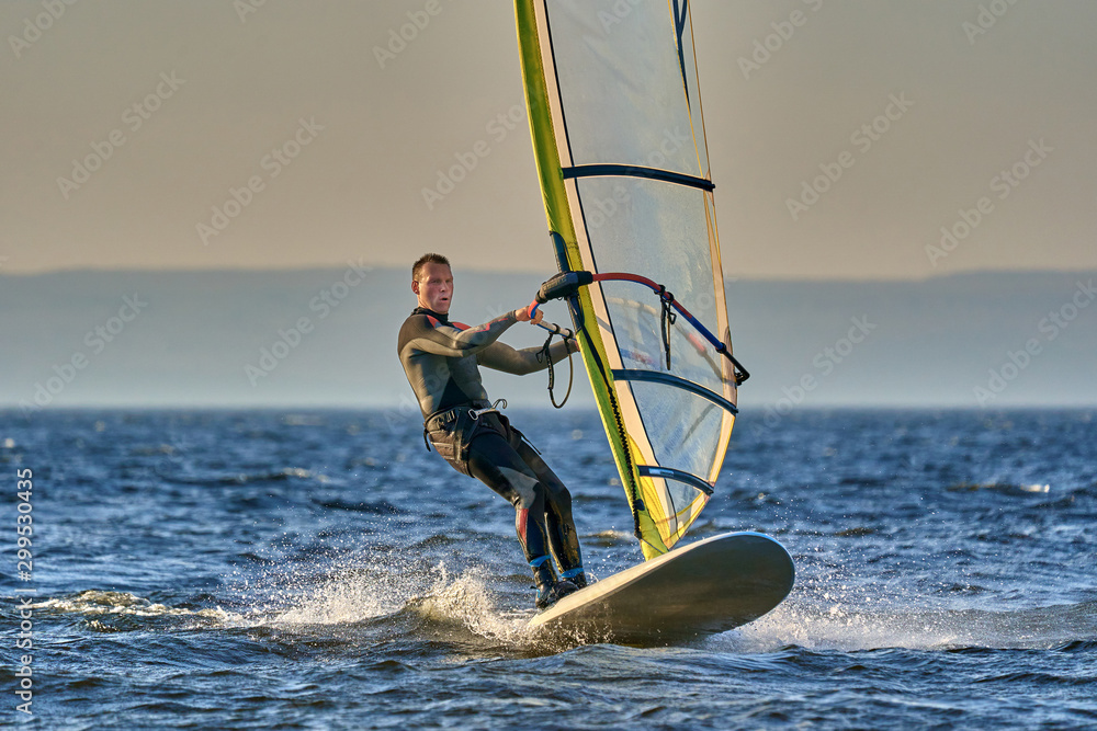 Naklejka premium A male athlete is interested in windsurfing. He moves on a Sailboard on a large lake on an autumn day.