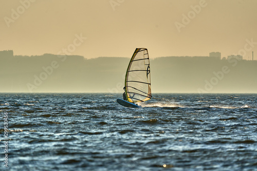 A male athlete is interested in windsurfing. He moves on a Sailboard on a large lake on an autumn day. © afefelov68