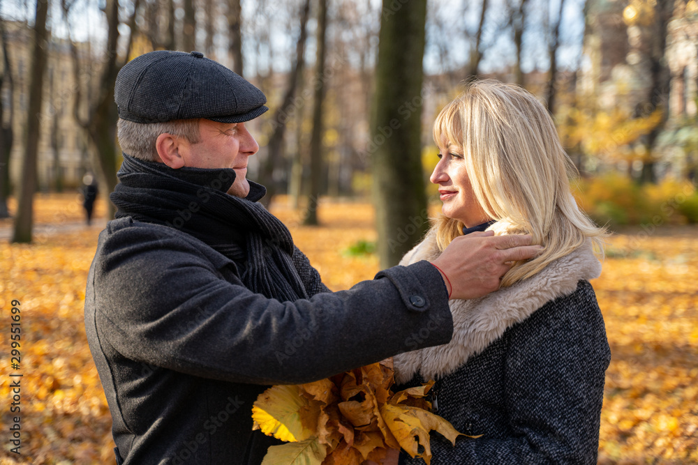 Fototapeta premium Handsome brunette middle-aged man carefully adjusts his hair. Happy blonde mature woman. Loving couple 45-50 years old are walking in park, looking at each other