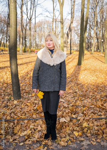 Happy blonde mature woman is standing in autumn park and thinking. Beautiful woman is relaxing in nature on sunny day. Portrait of middle aged woman smiling and daydreaming.