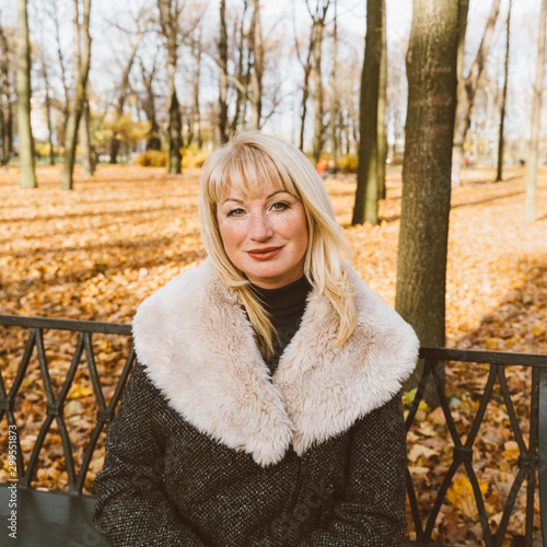 Happy blonde mature woman is sitting on bench in autumn park and thinking. Beautiful woman is relaxing in nature on sunny day. Portrait of middle aged woman smiling and daydreaming.