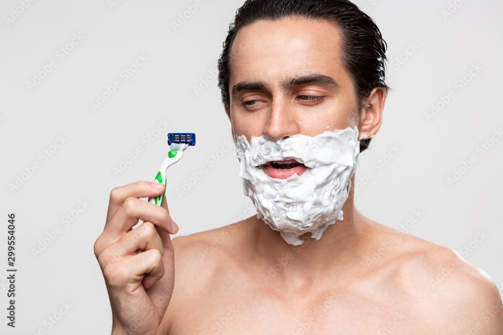Handsome man with mustache uses shaving stick and shaving foam to trim his beard standning bare isolated over white background. Concept of morning treatment and shaving.