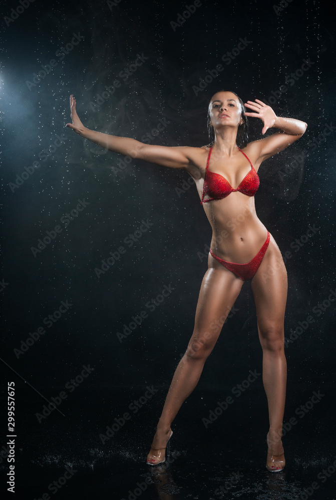 Beautiful wet muscular athletic girl wearing red underwear posing in scenic smoke and fog under falling water drops of rain on black background. Healthy smooth skin. Advertising and commercial design.
