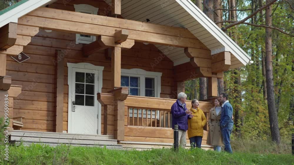 Wide shot of senior couple and their adult son with girlfriend standing near countryside wooden house and talking cheerfully. Two generation family spending weekend together Wide shot of senior couple and their adult son with girlfriend standing near countryside wooden house and talking cheerfully. Two generation family spending weekend together