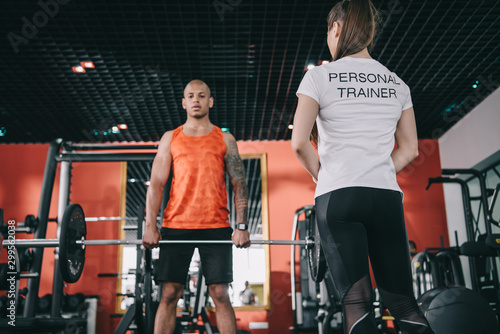 back view of personal trainer standing near african american athlete lifting barbell