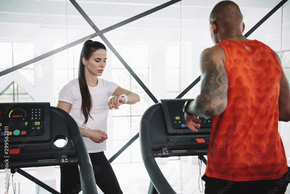 attentive trainer looking at fitness tracker while standing near african american sportsman running on treadmill