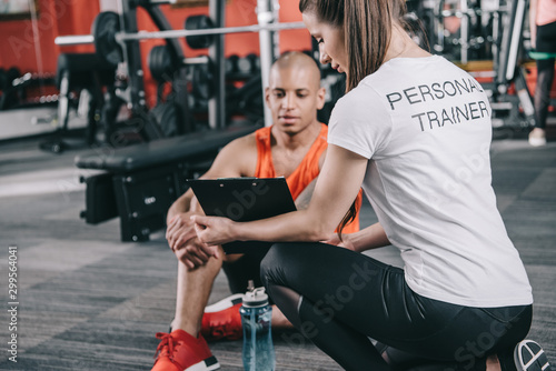 personal trainer showing clipboard to african american sportsman sitting on floor