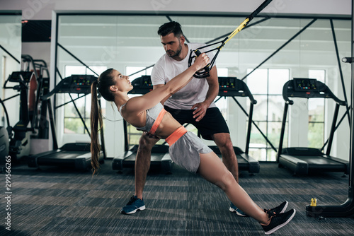 attentive trainer supporting young sportswoman pulling up on suspension trainer