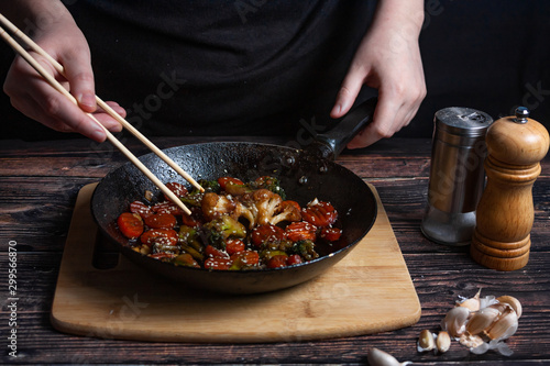 Chef at work. Hand holds a chopsticks under hot pan with fried vegetables, broccoli, cauliflower, carrots.