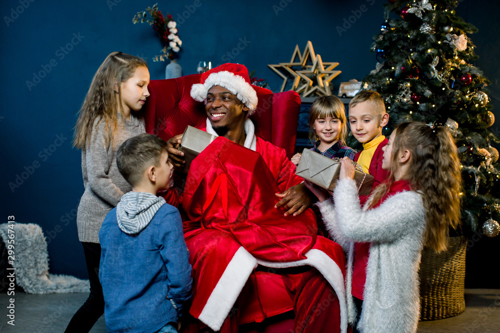 African Santa Claus is seated and gives presents to beautiful little children in a christmas-decorated room. African Santa Claus and happy little children on the background of the Christmas tree