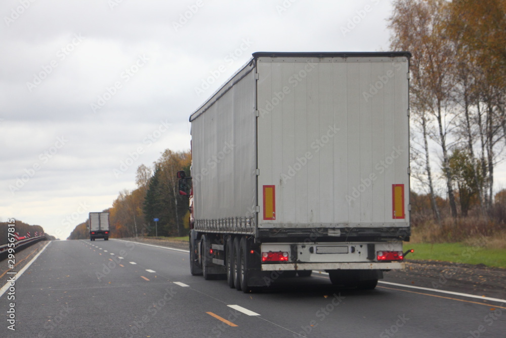 European white tree-axle semi truck with a semitrailer move on right lane autumn asphalt road, back view, transportation logistics European white tree-axle semi truck with a semitrailer move on right lane autumn asphalt road, back view, transportation logistics