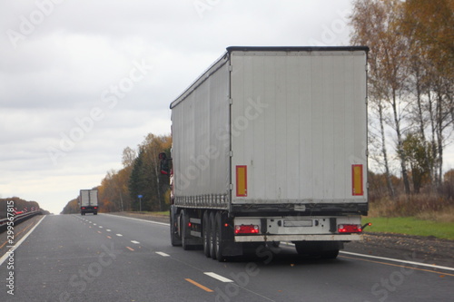 European white tree-axle semi truck with a semitrailer move on right lane autumn asphalt road, back view, transportation logistics