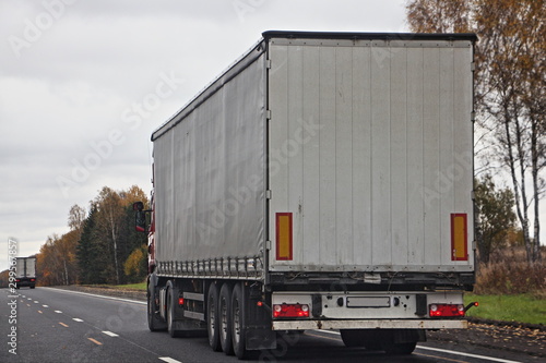 Euro white tree-axle semi truck with a semitrailer van drive on autumn asphalt road, close up rear side view, international cargo logistics business