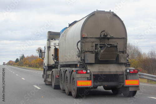 Tar truck with a round barrel semi trailer moving on a two-lane asphalted country road in autumn day, Logistics, hot liquid material road transportation, side rear view