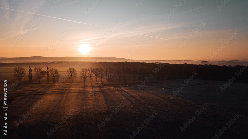Aerial view from above of a countryside landscape with beautiful sunset light on a winter evening with forzen meadows and mist hazy horizon. Winter autumn trees with long shadows in germany