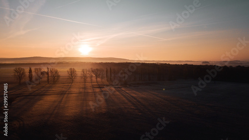Aerial view from above of a countryside landscape with beautiful sunset light on a winter evening with forzen meadows and mist hazy horizon. Winter autumn trees with long shadows in germany © Ricardo