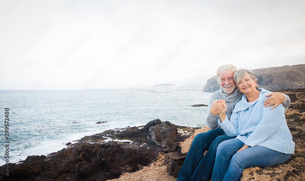 Couple of senior people sitting on the cliff of the ocean and looking at camera Hugging and smiling. Morning soon outside, ready for un healthy excursion. Vacation and happiness