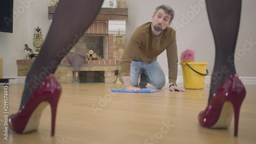 Portrait of adult Caucasian guy washing the floor and talking to his wife or girlfriend. Beautiful female legs in elegant red high-heel shoes on the foreground. Guilty henpecked doing housework. photo