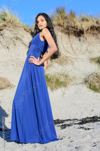 Beautiful model looking down at camera at the beach in Sligo in Ireland. Mixed race Portuguese Indian female model posing. Blue dress.  Black hair model. Irish coastline.