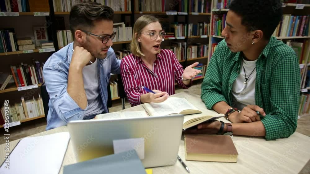 Three young students study in the school library, talking and using laptop for researching online. Three young students study in the school library, talking and using laptop for researching online.