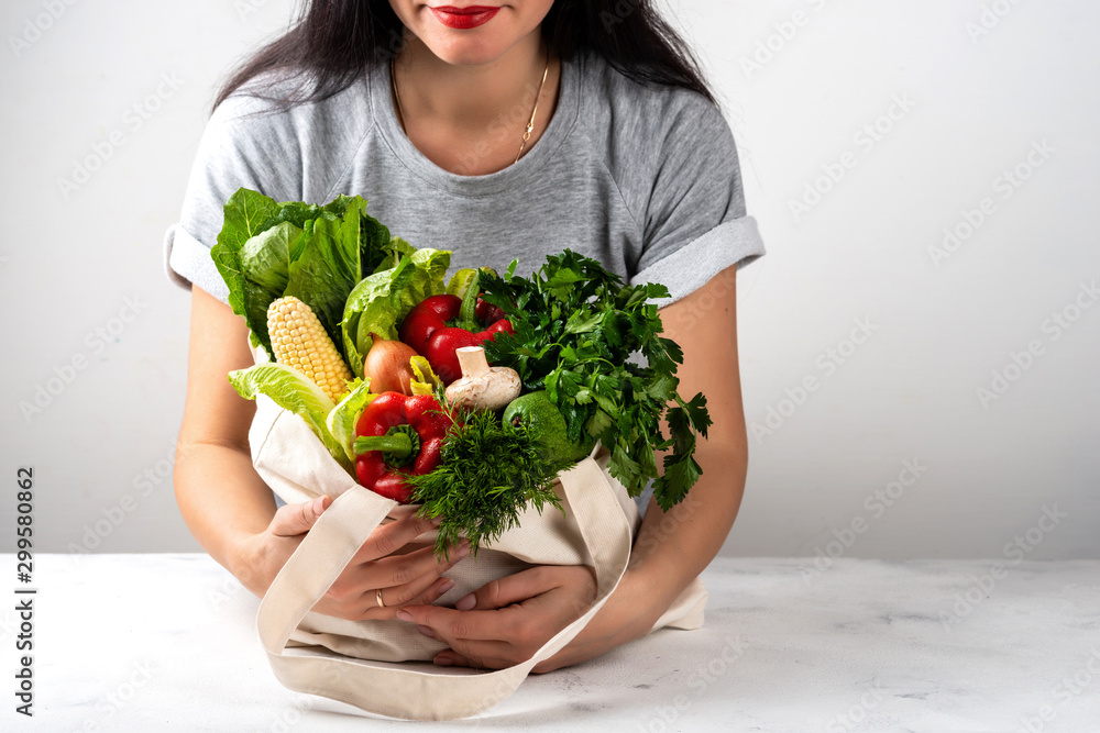 Woman holds a fabric eco bag full of different fresh vegetables in the bright kitchen. Purchases concept zero waste. Clean diet eating. Woman holds a fabric eco bag full of different fresh vegetables in the bright kitchen. Purchases concept zero waste. Clean diet eating.