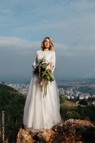 Beautiful elegant bride in lace wedding dress with long full skirt and long sleeves. She is holding a big bouquet of flowers. Nature, with city in the background. © Melika
