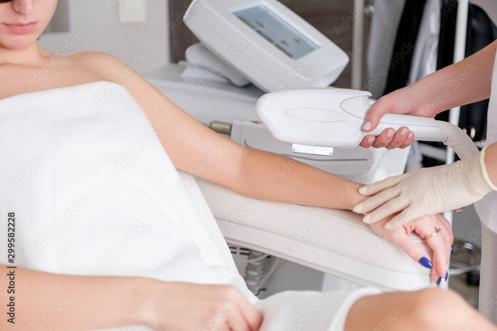 Cosmetologist's hands make photo-epilation using the apparatus to a young unidentified female client sitting in the chair of the beauty parlor. Body hair removal concept Cosmetologist's hands make photo-epilation using the apparatus to a young unidentified female client sitting in the chair of the beauty parlor. Body hair removal concept