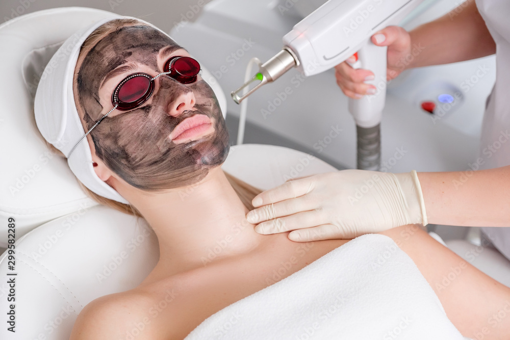 Naklejka premium Close-up of a cosmetologist's hand making a hardware carbon peeling by a young pretty customer woman in safety glasses and a headdress lying on a couch in a beauty salon. Facial cleansing concept