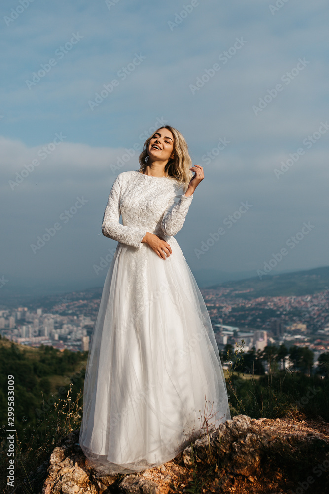 Beautiful elegant bride in lace wedding dress with long full skirt and long sleeves. Pretty girl in white. Nature, with city in the background. Beautiful elegant bride in lace wedding dress with long full skirt and long sleeves. Pretty girl in white. Nature, with city in the background.