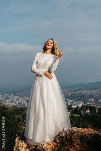 Beautiful elegant bride in lace wedding dress with long full skirt and long sleeves. Pretty girl in white. Nature, with city in the background. © Melika
