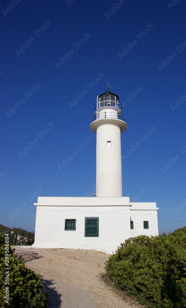 Old stone built lighthouse of Lefkada island south cape at summer, clear deep blue sky at background. A path through shrubs and bushes at the foreground