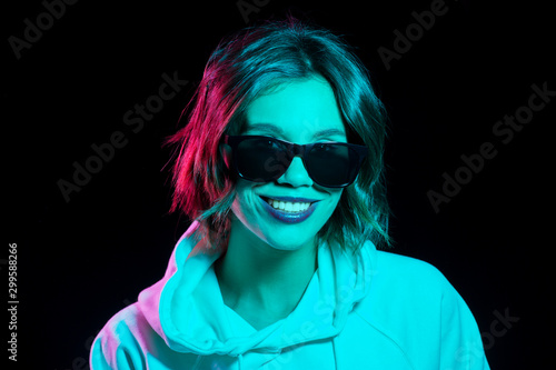 nightlife, fashion and people concept - happy young woman wearing hoodie in neon lights over black background