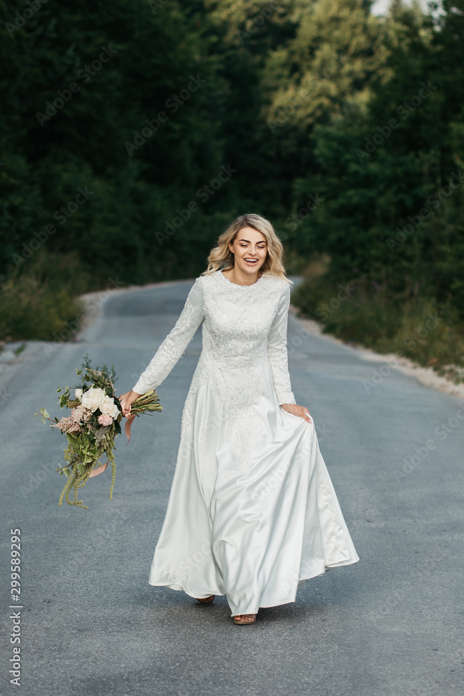 Beautiful elegant bride in lace wedding dress with long full skirt and long sleeves. She is holding a big bouquet of flowers. Outdoors, on the road. Beautiful elegant bride in lace wedding dress with long full skirt and long sleeves. She is holding a big bouquet of flowers. Outdoors, on the road.