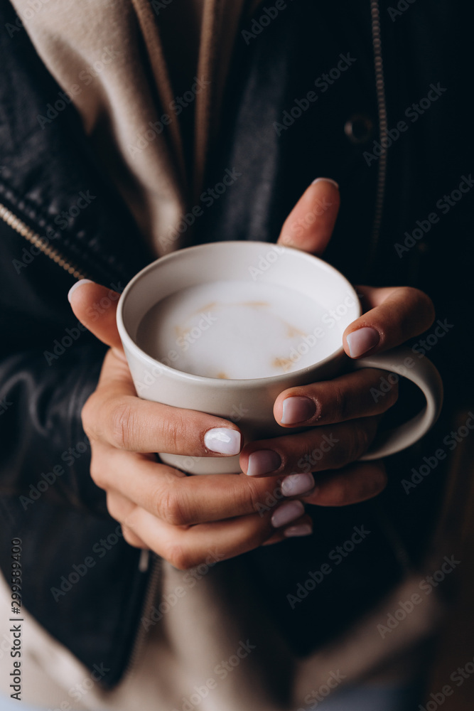 close up female dressed in autumn clothes holds in two hands a big cup of hot coffee with milk standing on the street. The girl warms the cup and enjoys aromatic coffee close up female dressed in autumn clothes holds in two hands a big cup of hot coffee with milk standing on the street. The girl warms the cup and enjoys aromatic coffee