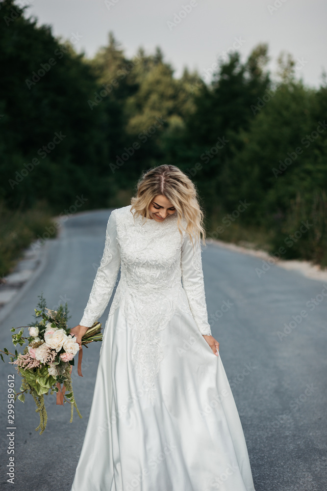 Beautiful elegant bride in lace wedding dress with long full skirt and long sleeves. She is holding a big bouquet of flowers. Outdoors, on the road. Beautiful elegant bride in lace wedding dress with long full skirt and long sleeves. She is holding a big bouquet of flowers. Outdoors, on the road.