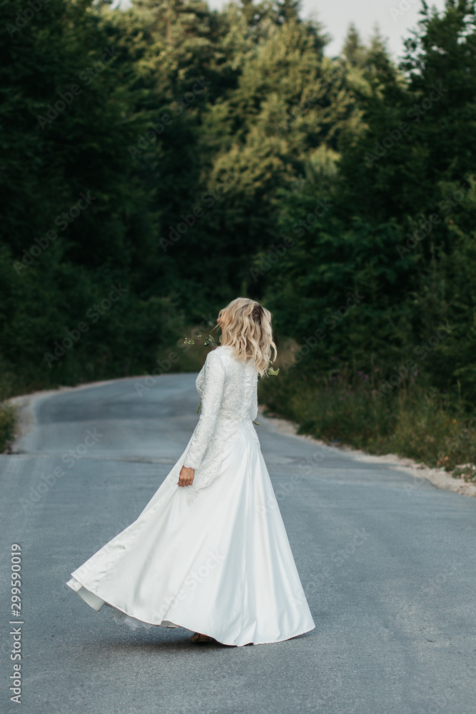 Beautiful elegant bride in lace wedding dress with long full skirt and long sleeves. She is holding a big bouquet of flowers. Outdoors, on the road.