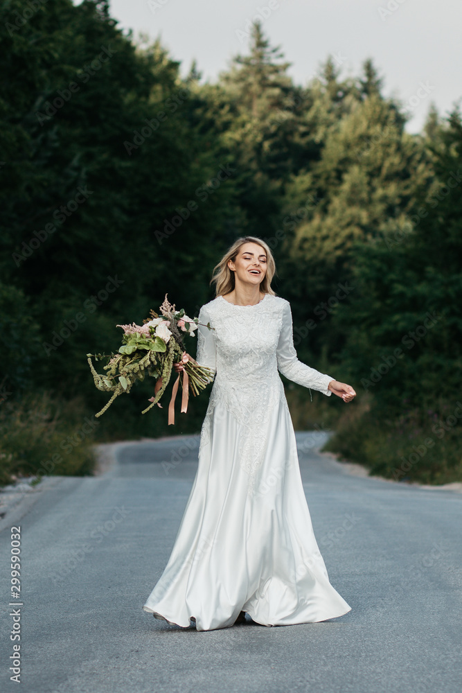 Beautiful elegant bride in lace wedding dress with long full skirt and long sleeves. She is holding a big bouquet of flowers. Outdoors, on the road. Beautiful elegant bride in lace wedding dress with long full skirt and long sleeves. She is holding a big bouquet of flowers. Outdoors, on the road.