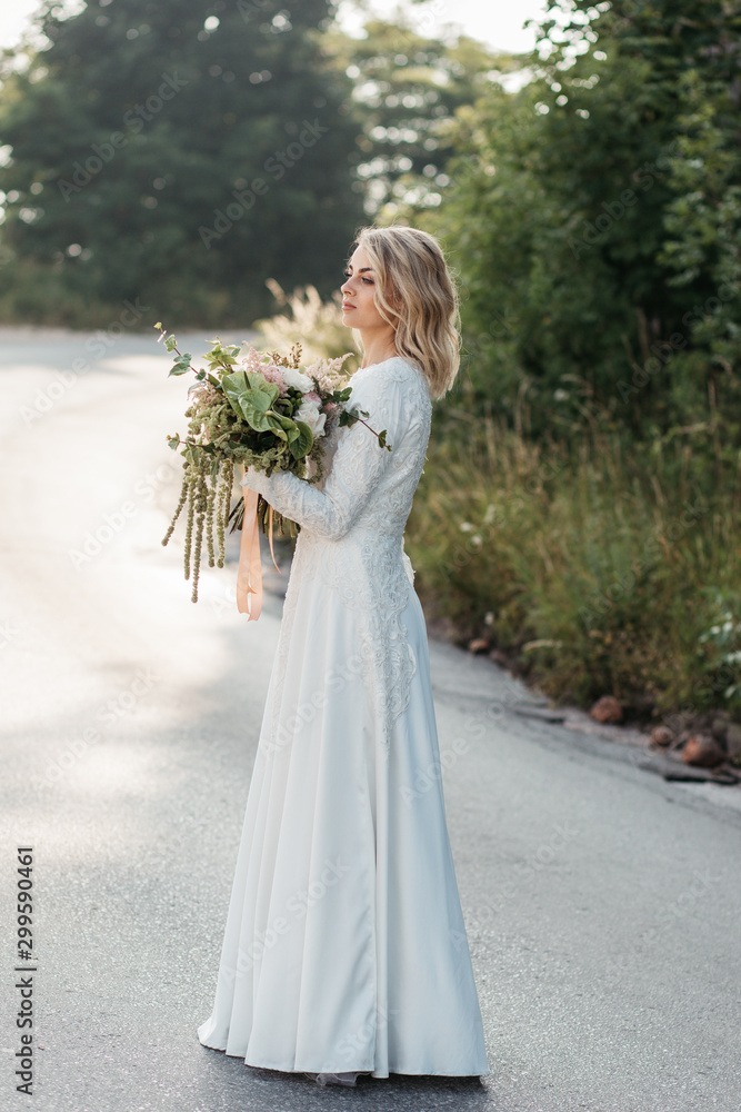 Beautiful elegant bride in lace wedding dress with long full skirt and long sleeves. She is holding a big bouquet of flowers. Outdoors, on the road. Beautiful elegant bride in lace wedding dress with long full skirt and long sleeves. She is holding a big bouquet of flowers. Outdoors, on the road.
